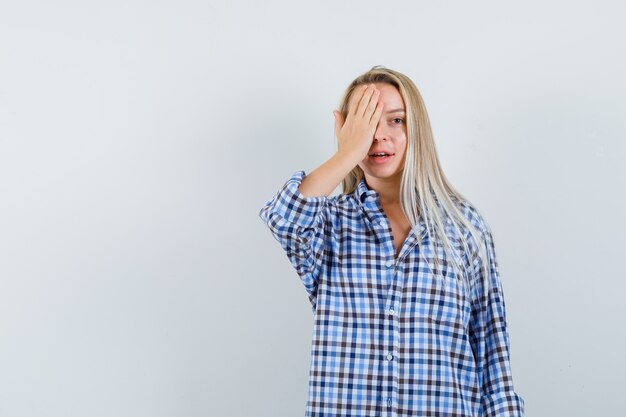 Blonde Dame, die Hand auf einem Auge im karierten Hemd hält und positiv schaut