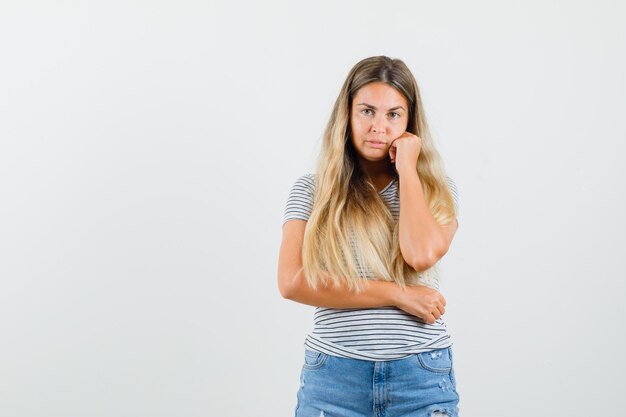 Blonde Dame, die Hand auf ihr Gesicht im T-Shirt setzt und gelangweilt, Vorderansicht schaut.