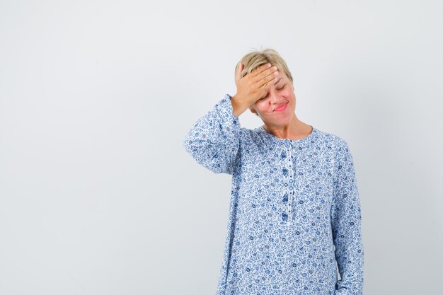 Blonde Dame, die Hand auf ihrer Stirn in gemusterter Bluse hält und müde, Vorderansicht schaut.