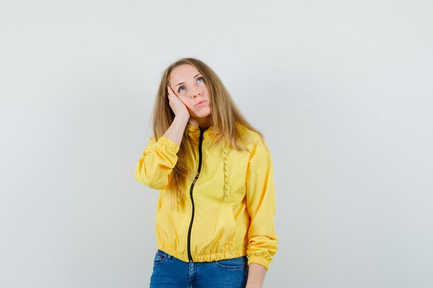 Blonde Dame, die Hand auf Wange in Jacke, Jeans hält und nachdenklich aussieht,