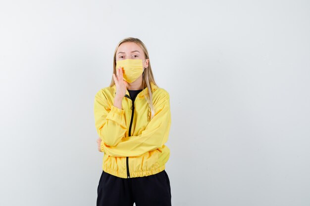 Blonde Dame, die Hand nahe Mund hält, um Geheimnis im Trainingsanzug, in der Maske, in der Vorderansicht zu verraten.