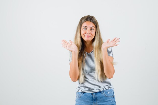 Blonde Dame, die Handflächen in Übergabegeste in gestreiftem T-Shirt, Jeans zeigt und verwirrt aussieht. Vorderansicht.