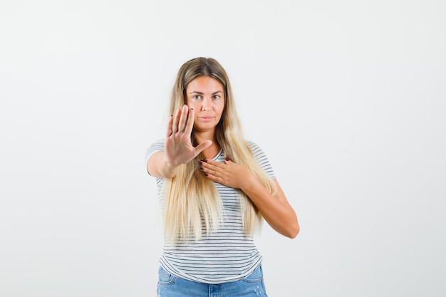 Blonde Dame, die ihre Hand auf ablehnende Weise im T-Shirt hebt und ernst aussieht. Vorderansicht.