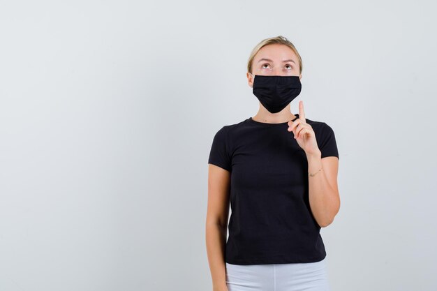 Blonde Dame, die in schwarzem T-Shirt, schwarzer Maske nach oben zeigt und konzentriert aussieht