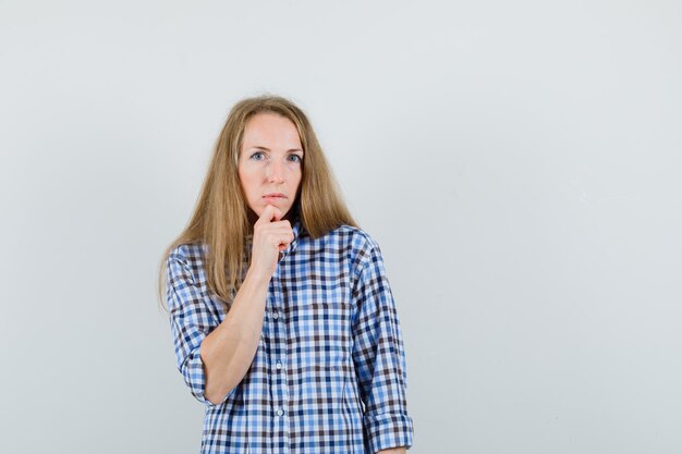 Blonde Dame, die Kinn auf Hand im Hemd stützt und zögernd aussieht.