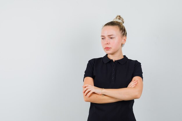 Blonde Dame, die mit verschränkten Armen im schwarzen T-Shirt steht und verärgert schaut, Vorderansicht.