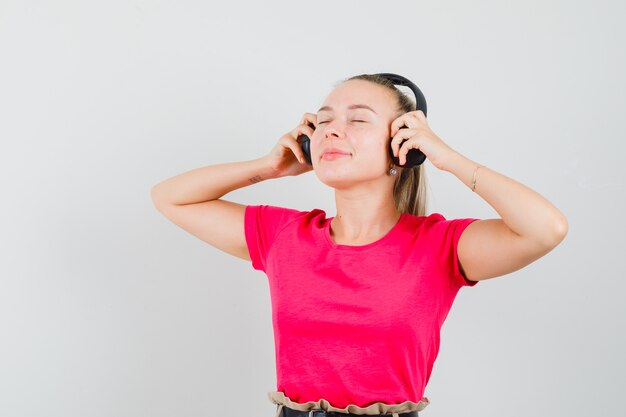 Blonde Dame, die Musik mit Kopfhörern im rosa T-Shirt genießt und entzückte Vorderansicht schaut.