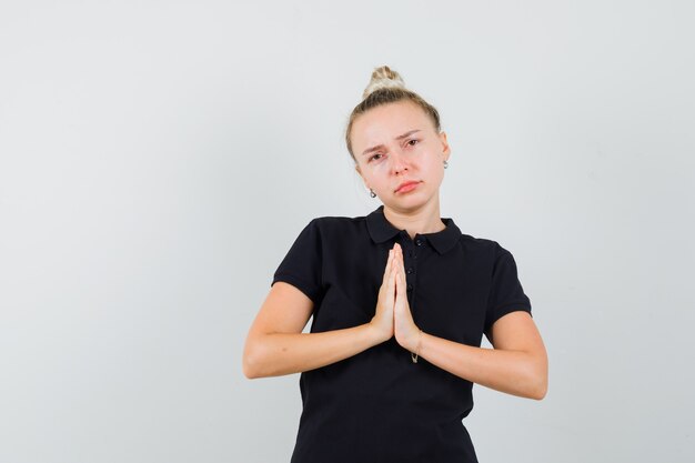 Blonde Dame, die namaste Geste im schwarzen T-Shirt zeigt und traurig, Vorderansicht schaut.