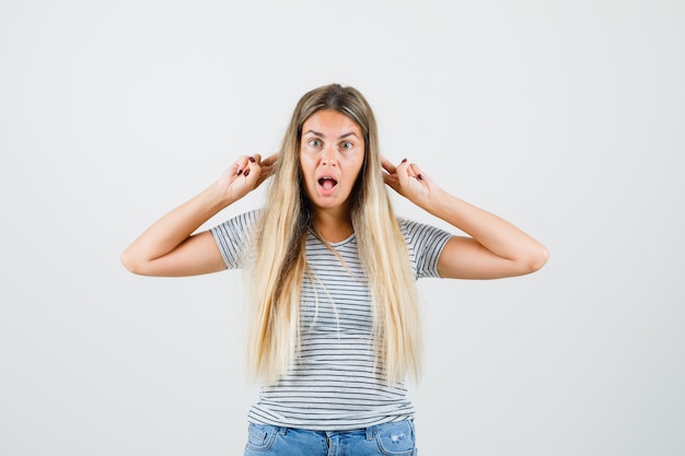 Blonde Dame, die Ohren mit den Fingern im gestreiften T-Shirt, in den Jeans und in der lustigen Vorderansicht schiebt.