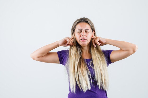 Blonde Dame, die Ohren mit den Fingern im violetten T-Shirt verstopft und genervt aussieht. Vorderansicht.