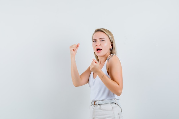 Blonde Dame, die Siegergeste im Unterhemd, in den Hosen zeigt und glückliches, Vorderansicht schaut.