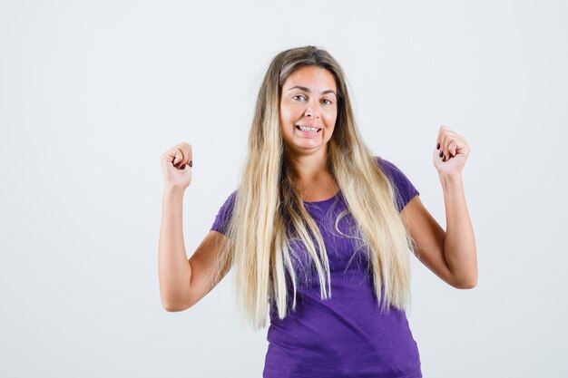 Blonde Dame, die Siegergeste im violetten T-Shirt zeigt und glückselig aussieht. Vorderansicht.