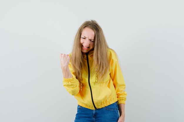 Blonde Dame, die Siegergeste in Jacke, Jeans zeigt und glücklich schaut.