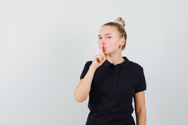 Blonde Dame, die Stille Geste im schwarzen T-Shirt zeigt und vorsichtig schaut. Vorderansicht.