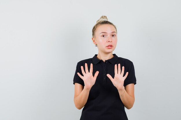 Blonde Dame, die Stoppgeste im schwarzen T-Shirt zeigt und ängstlich aussieht. Vorderansicht.