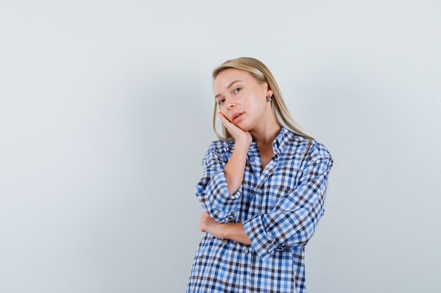 Blonde Dame, die Wange auf erhabener Handfläche im karierten Hemd lehnt und niedlich aussieht