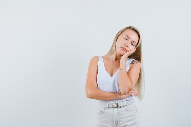 Blonde Dame, die Wange auf erhabener Handfläche im Unterhemd, in der Hose lehnt und schläfrig aussieht