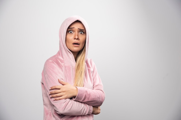 Blonde Dame im rosa Sweatshirt macht erschrockenes und verängstigtes Gesicht im Hoodie.