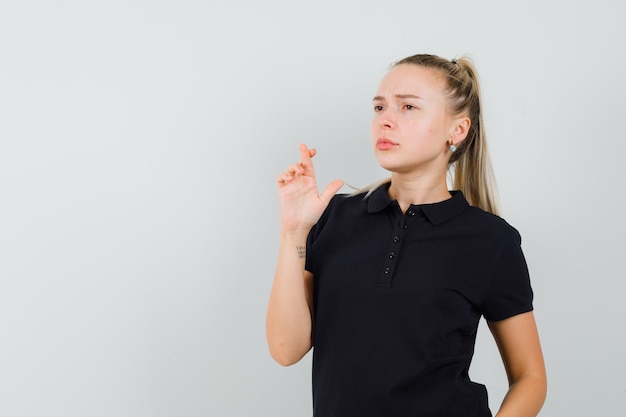 Blonde Dame im schwarzen T-Shirt, das Daumen drückt und nachdenklich aussieht, Vorderansicht.