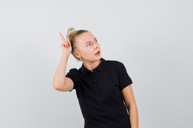 Blonde Dame im schwarzen T-Shirt, das Finger über Kopf hält und neugierige Vorderansicht schaut.