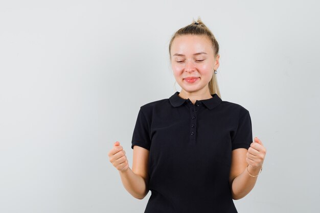 Blonde Dame im schwarzen T-Shirt hält geballte Fäuste und schaut freudig, Vorderansicht.