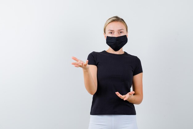 Blonde Dame im schwarzen T-Shirt, schwarze Maske, die Hände in fragender Geste isoliert hält keeping