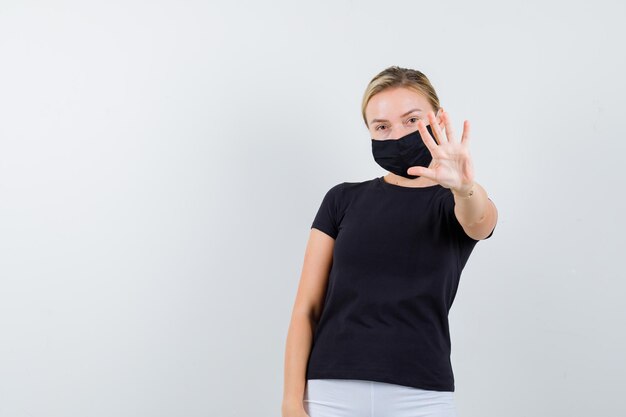 Blonde Dame im schwarzen T-Shirt, schwarze Maske mit Stopp-Geste und selbstbewusstem Blick