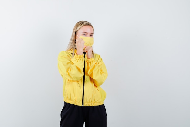 Blonde Dame im Trainingsanzug, Maske stehend in Kampfhaltung und selbstbewusst aussehend, Vorderansicht.