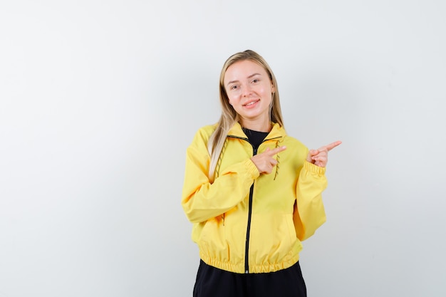 Blonde Dame im Trainingsanzug zeigt nach rechts und sieht fröhlich aus, Vorderansicht.