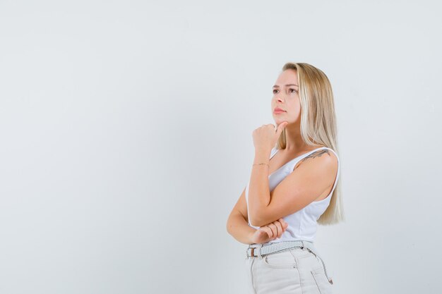 Blonde Dame im Unterhemd, Hose stützt das Kinn auf die Hand und sieht nachdenklich aus.