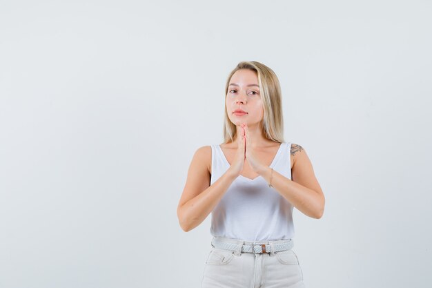 Blonde Dame im Unterhemd, Hose zeigt namaste Geste und sieht hoffnungsvoll aus