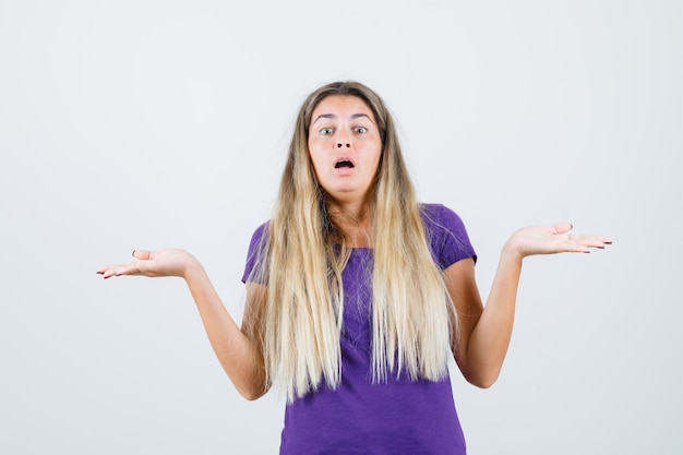 Blonde Dame im violetten T-Shirt, das Schuppengeste zeigt und verwirrt, Vorderansicht schaut.