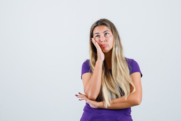 Blonde Dame im violetten T-Shirt hält Handfläche auf Wange und sieht nachdenklich, Vorderansicht.