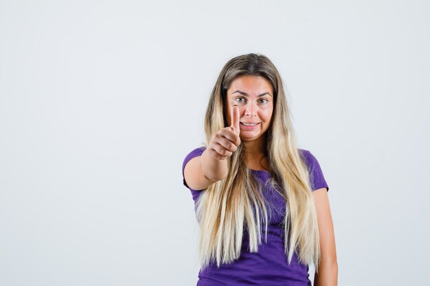 Blonde Dame im violetten T-Shirt zeigt Daumen hoch und sieht fröhlich aus, Vorderansicht.