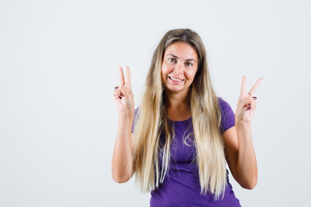 Blonde Dame im violetten T-Shirt zeigt Siegesgeste und schaut fröhlich, Vorderansicht.