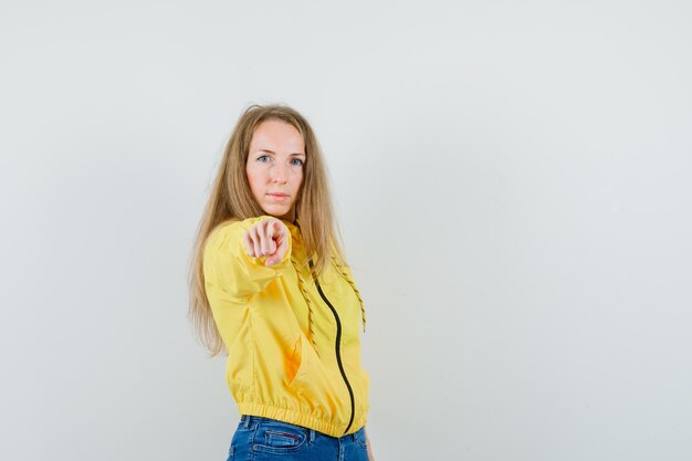 Blonde Dame in Jacke, Jeans zeigt auf Kamera und sieht selbstbewusst aus,