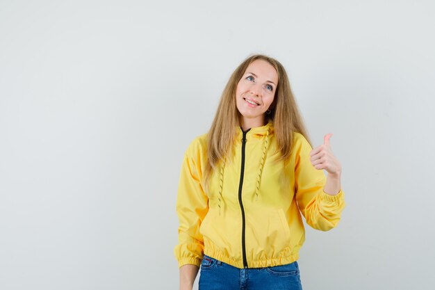 Blonde Dame in Jacke, Jeans zeigt Daumen hoch und sieht fröhlich aus,