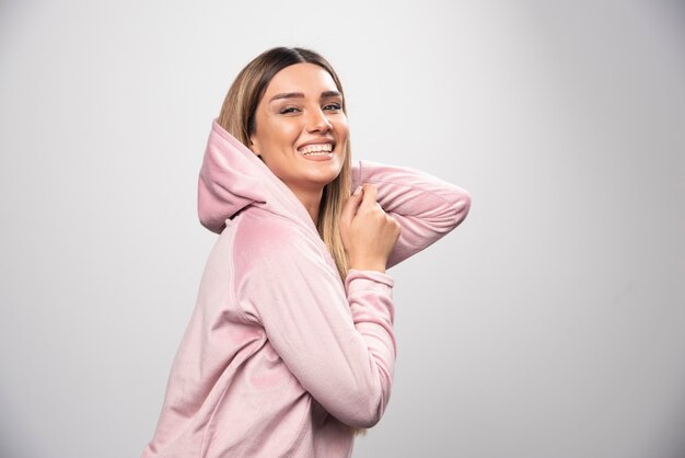 Blonde Dame in rosa Sweatshirt macht freudige und positive Posen, indem sie Hoodie an ihrem Kopf trägt.