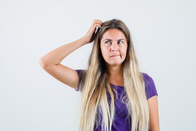 Blonde Dame kratzt den Kopf im violetten T-Shirt und sieht nachdenklich aus, Vorderansicht.