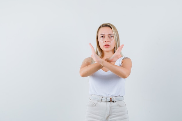 Blonde Dame zeigt Ablehnungsgeste im Unterhemd, in der Hose und sieht ernst aus