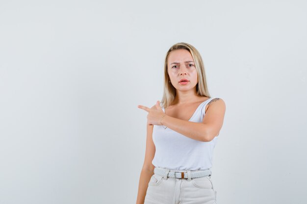 Blonde Dame zeigt auf die linke Seite in Unterhemd, Hose und sieht ernst aus, Vorderansicht.