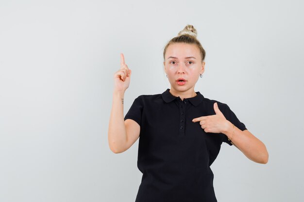 Blonde Dame zeigt auf sich selbst mit dem Finger im schwarzen T-Shirt und sieht verwirrt aus. Vorderansicht.