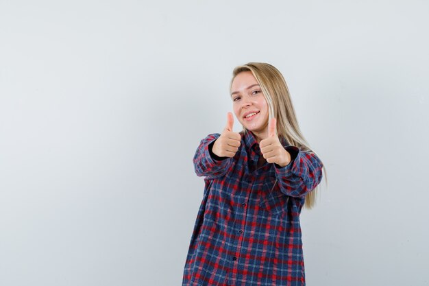 Blonde Dame zeigt Daumen hoch im Freizeithemd und sieht fröhlich aus. Vorderansicht.