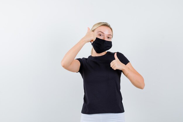 Blonde Dame zeigt Daumen hoch in schwarzem T-Shirt, schwarzer Maske und sieht zufrieden aus