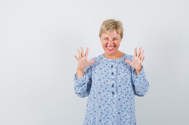 Blonde Dame zeigt ihre offenen Handflächen in gemusterter Bluse und sieht lustig aus. Vorderansicht. Platz für Text