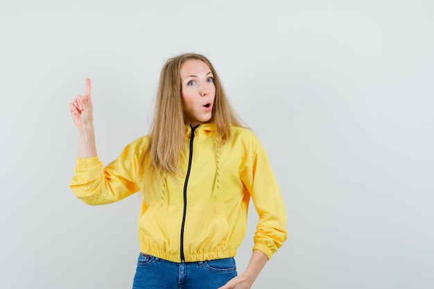 Blonde Dame zeigt in Jacke, Jeans und sieht selbstbewusst aus.