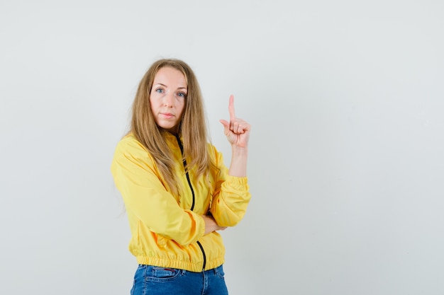 Blonde Dame zeigt in Jacke, Jeans und sieht selbstbewusst aus.