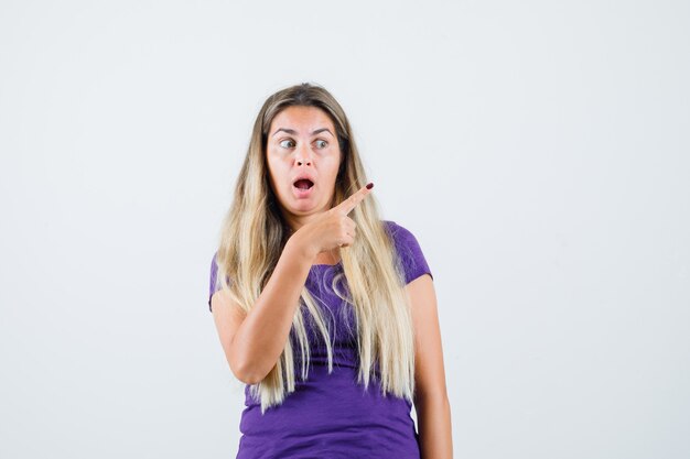 Blonde Dame zeigt in violettem T-Shirt weg und sieht verwundert aus. Vorderansicht.