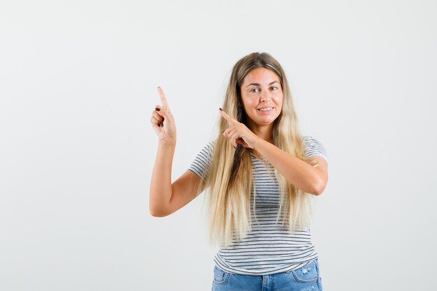 Blonde Dame zeigt nach oben, während sie im T-Shirt lächelt und froh aussieht. Vorderansicht.