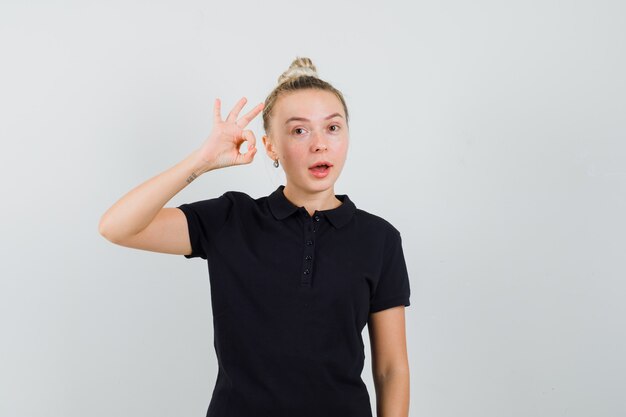 Blonde Dame zeigt ok Zeichen im schwarzen T-Shirt und schaut selbstbewusst, Vorderansicht.